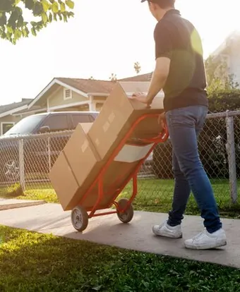 A man pulling a hand truck loaded with boxes along a pavement past houses with yards