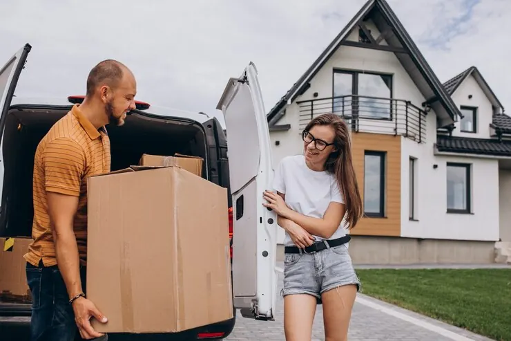 A man unloading a large box from a van outside a house while the woman moving in looks on